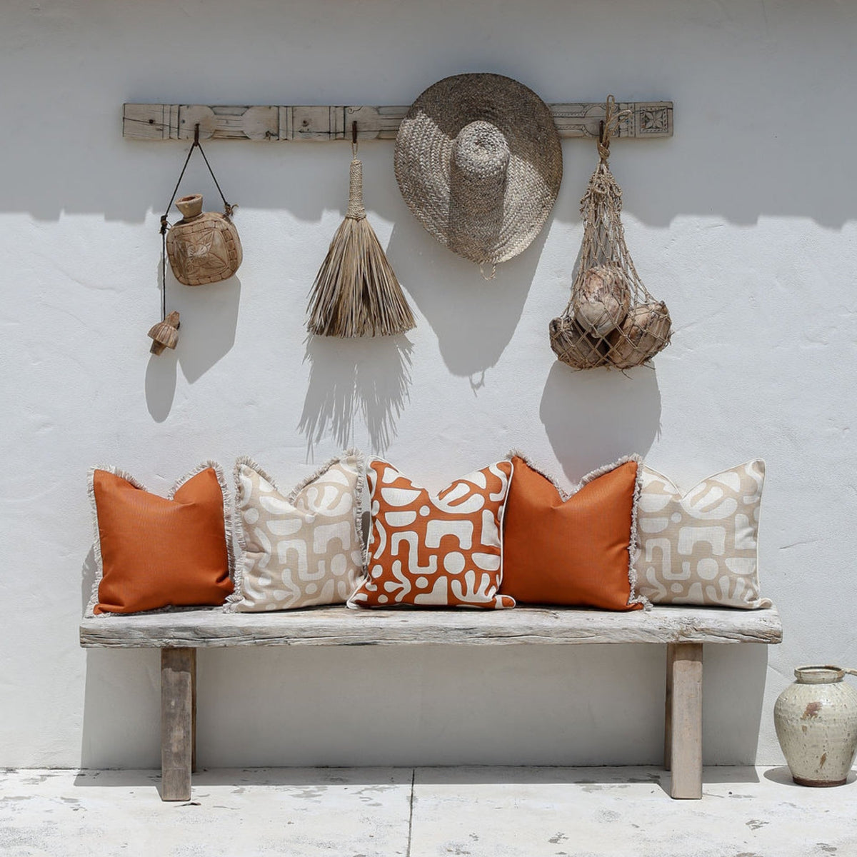 A rustic wooden bench with six decorative throw pillows in terracotta orange, cream, and orange-and-beige patterns, set against a white stucco wall with a wooden rack holding a straw hat and woven bags.