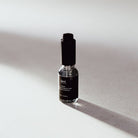 A small glass bottle with a black cap and black label, standing on a light gray surface with a strong diagonal shadow. The label shows the brand name 'kerri'.
