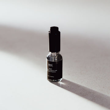 A small glass bottle with a black cap and black label, standing on a light gray surface with a strong diagonal shadow. The label shows the brand name 'kerri'.