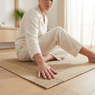 A woman in a cream-colored linen pajama set sits cross-legged on a natural jute yoga mat on a light wood floor in a bright, modern room.