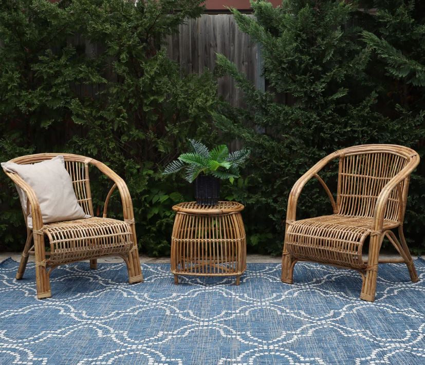 Two wicker chairs with a small table on a blue patterned rug against a green bush background