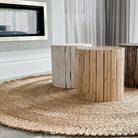 Three cylindrical wooden stools in light, medium, and dark natural wood tones arranged on a large round woven jute rug in a modern living room setting.