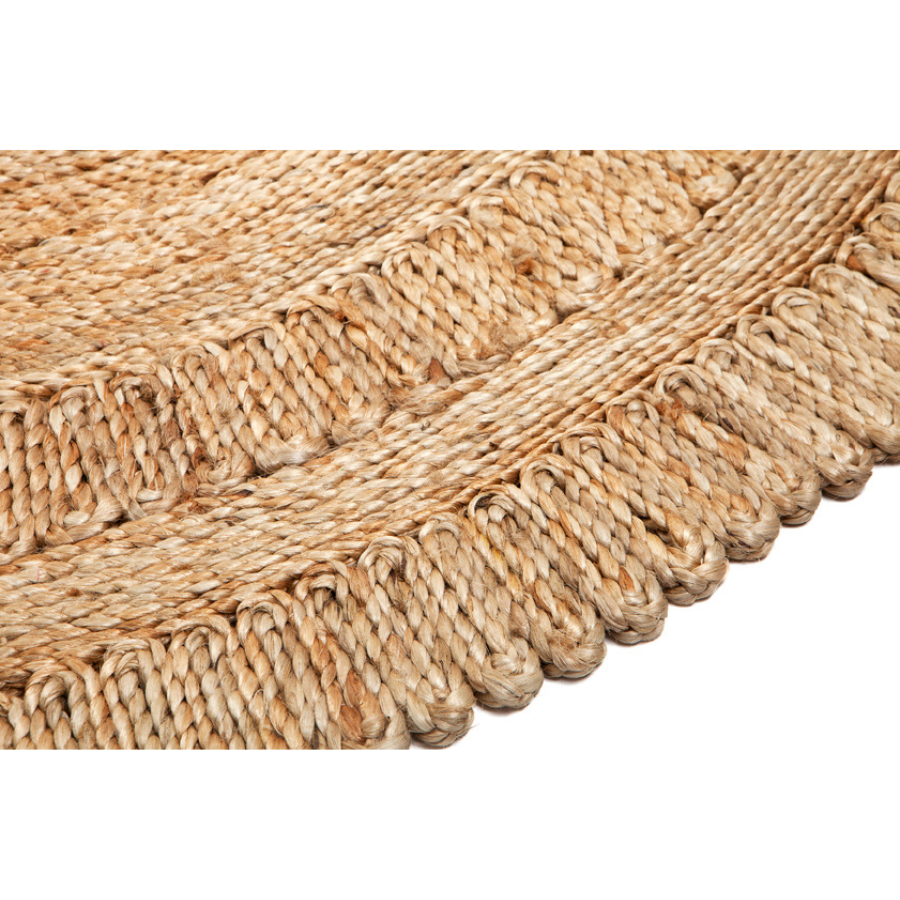 Close-up of a woven natural fiber product, likely a rug or mat, showing a braided texture and scalloped fringe edge in warm tan and beige tones against a white background.