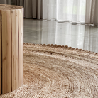 A close-up of a round natural jute rug with a braided texture and scalloped edge, shown in a modern room setting with a light wood cylindrical side table and sheer curtains in the background.