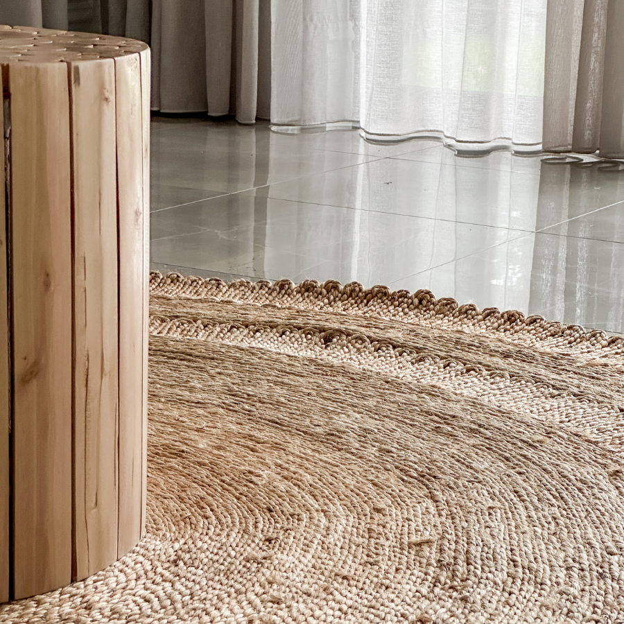 A close-up of a round natural jute rug with a braided texture and scalloped edge, shown in a modern room setting with a light wood cylindrical side table and sheer curtains in the background.