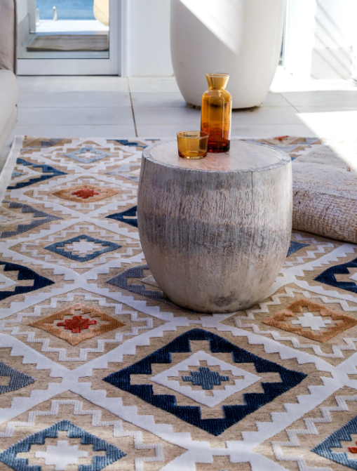 Decorative stone table with a glass and vase on a patterned rug in a bright room.