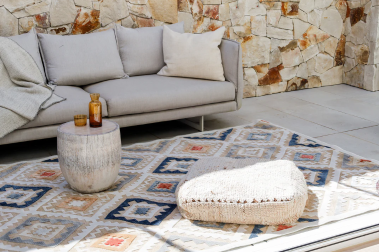 Gray sofa with cushions and a small table on a patterned rug against a stone wall.