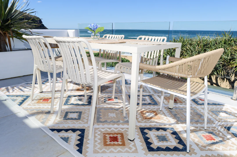 Outdoor dining set with table and chairs on a patterned rug by the sea.