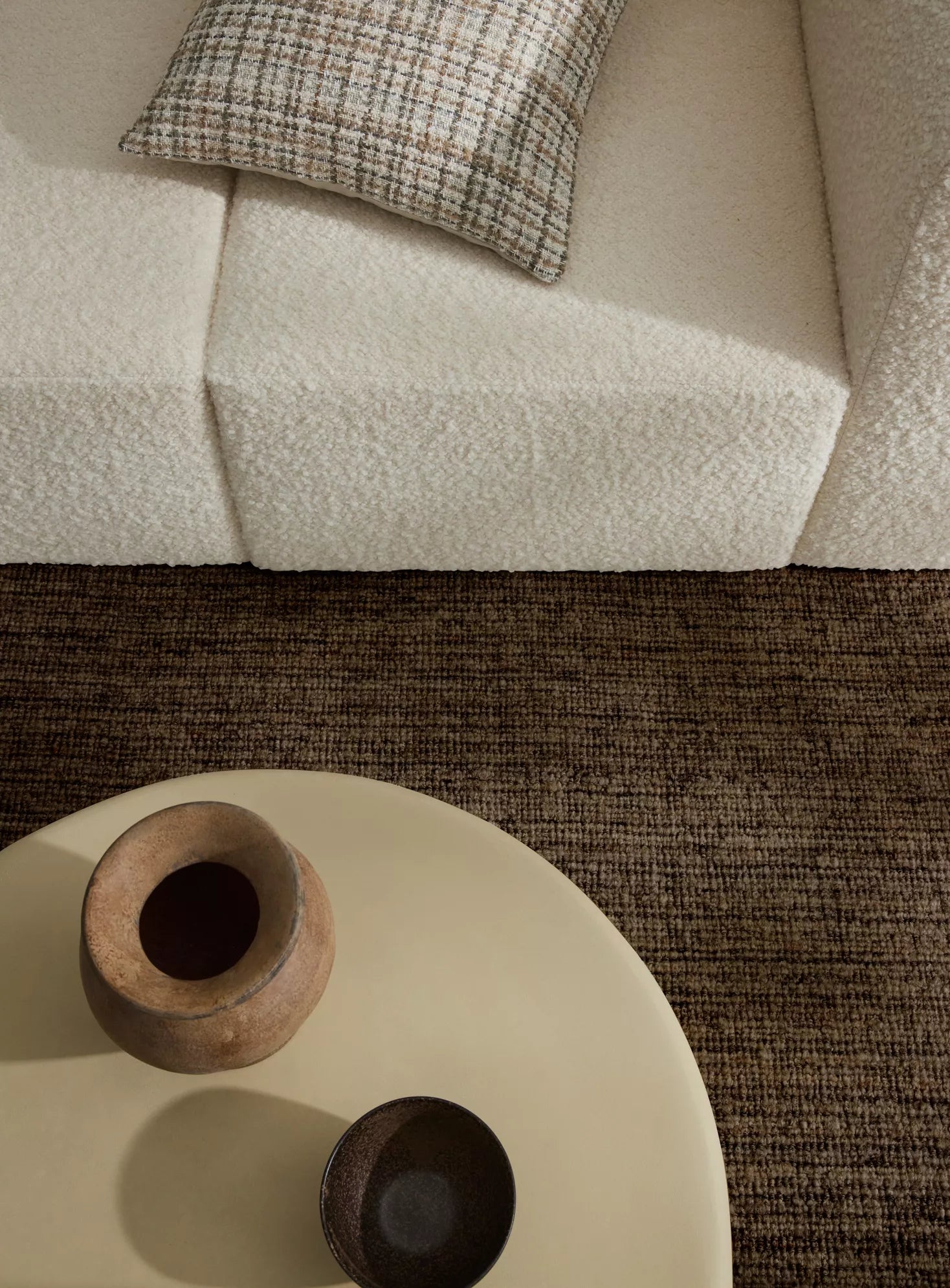 Round beige coffee table with wooden vase and bowl on a textured brown rug, next to a beige sofa with a patterned pillow.
