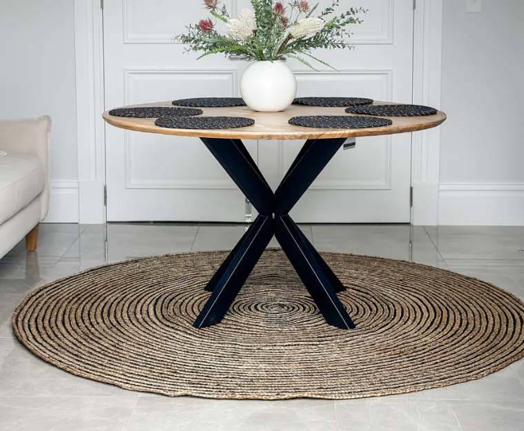 Round wooden table with black metal legs on a woven rug in a room.