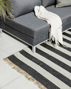 A modern gray sectional sofa with a white throw blanket, styled on a black and off-white striped woven area rug with fringed edges over a light tile floor.