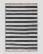 A rectangular woven rug with horizontal stripes in dark charcoal gray and off-white, finished with fringed edges on all sides, shown flat against a light gray background.