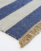 Close-up of a woven rug corner showing diagonal stripes in blue and beige with a fringed edge on a white surface.
