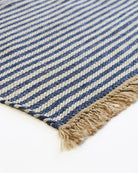 Close-up of a woven rug corner showing diagonal navy and cream stripes with a natural beige fringed edge on a white background.