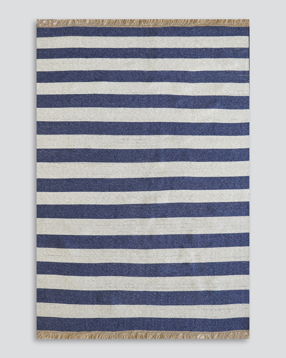 A rectangular woven rug with horizontal navy blue and off-white stripes, finished with natural jute fringe on the top and bottom edges, shown flat against a light gray background.