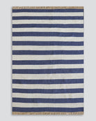 A rectangular woven rug with horizontal navy blue and off-white stripes, finished with natural jute fringe on the top and bottom edges, shown flat against a light gray background.