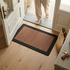 Brown doormat with black border on wooden floor, person entering through glass door.